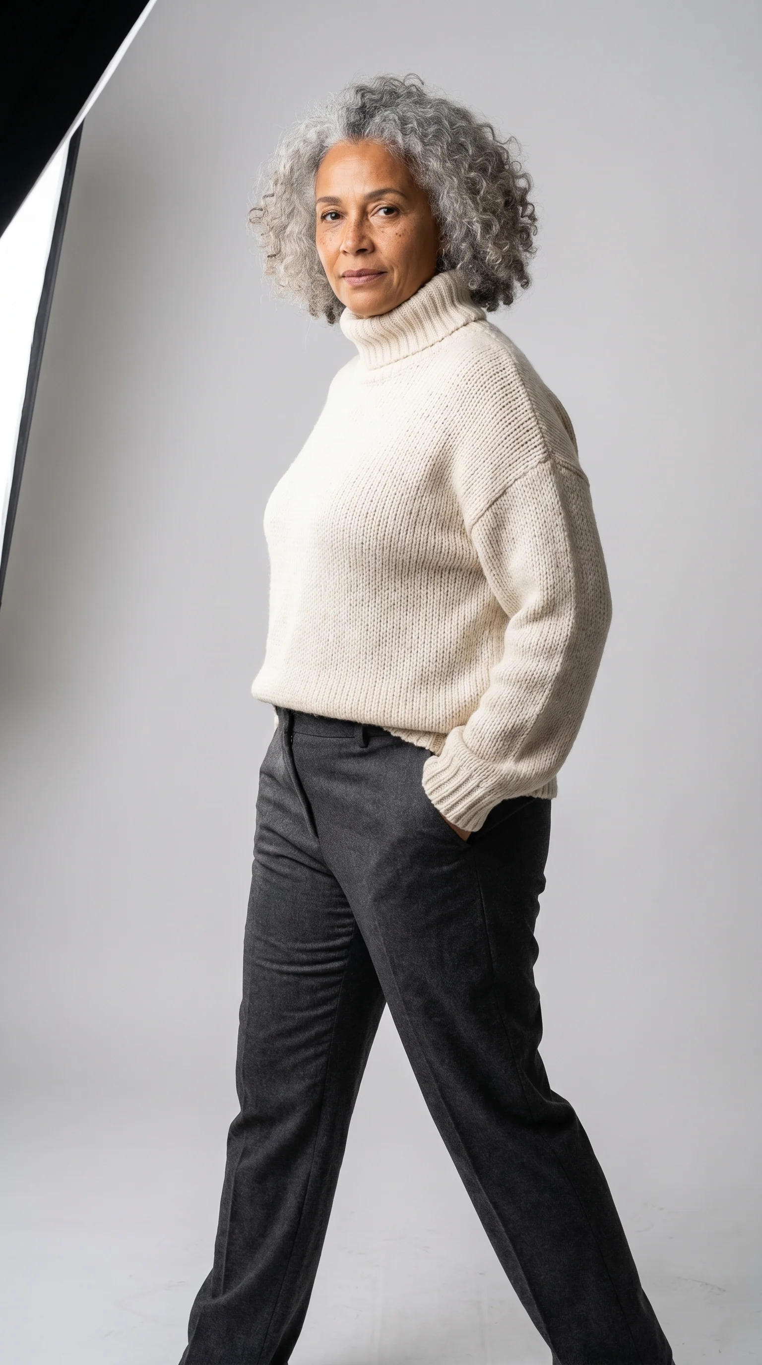 Modelo de uns 55 anos de cabelo grisalho cacheado, tricô creme oversized e calça de alfaiataria, fundo cinza de estúdio, trama do tecido visível.