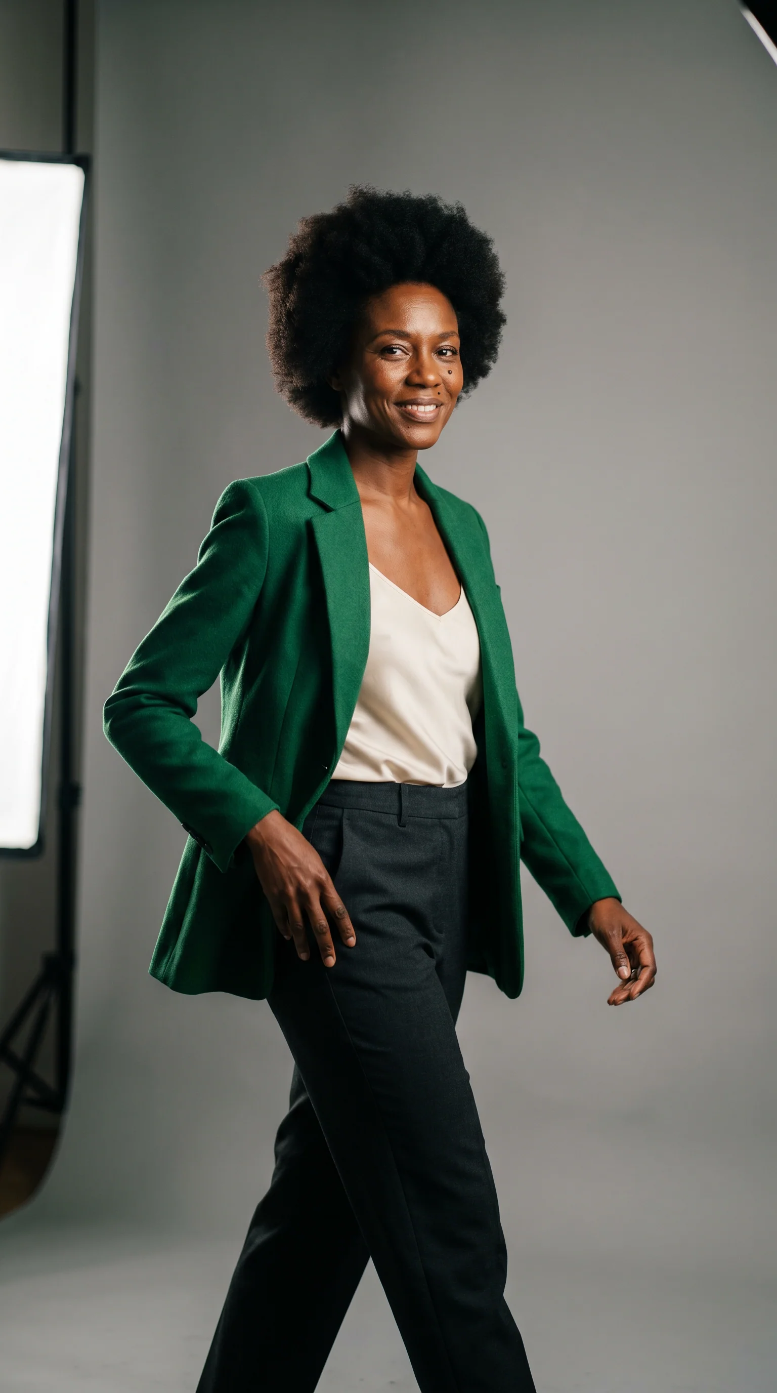 Mulher negra com afro curto e blazer verde-esmeralda, corpo inteiro num cyclorama cinza de estúdio, em meio passo com a luz de estúdio visível no olho.