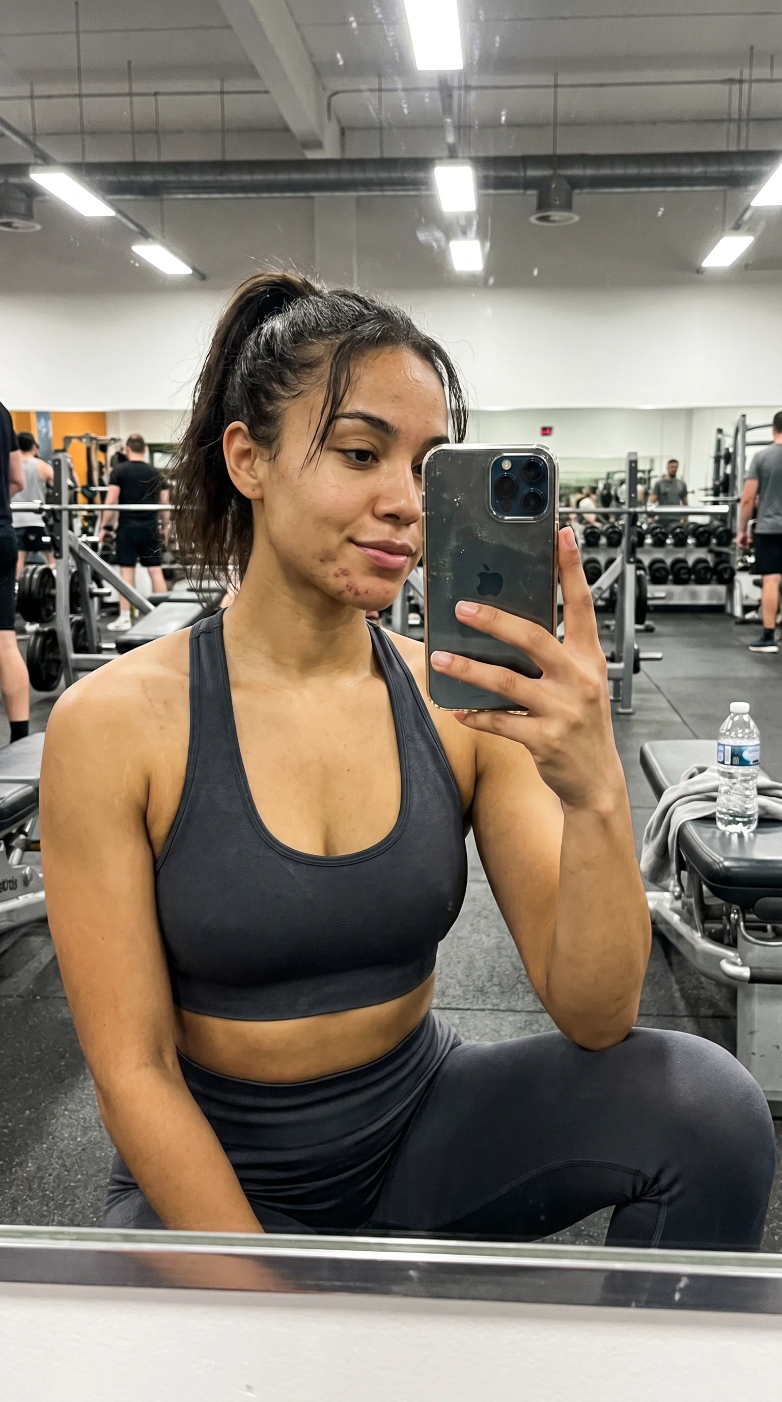 Selfie de academia: top esportivo escuro, academia real ao fundo com gente treinando, marcas de acne no queixo, celular refletido no espelho.